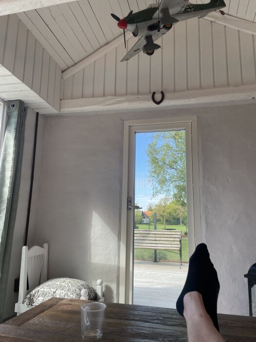 Utsikt från ett uterum med fot på träbord, glas, modellflygplan i taket, hästsko på vägg och utsikt mot trädgård genom dörr.