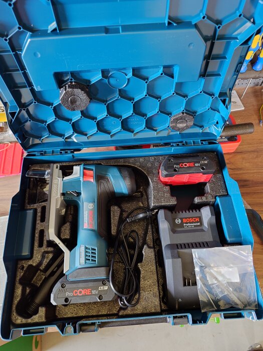 Blå förvaringslåda med ett Bosch elverktyg, laddare och batteri inuti, tillsammans med plastpåse med borrar.