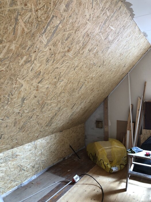 Snedvägg i sovrum med OSB-skivor, golv med träplankor. Verktyg på golvet, en plastförpackning med isolering och en träpall syns. Renovationsprojekt.