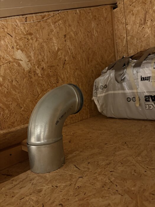 Ventilationsrör i plåt ovanpå träskiva i ett utrymme med spånskivor på väggarna, bredvid en säck isoleringsmaterial.