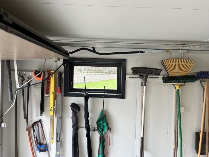 Verktyg och sopborstar hänger organiserat på en vägg i ett garage, med ett litet fönster som släpper in ljus ovanför.