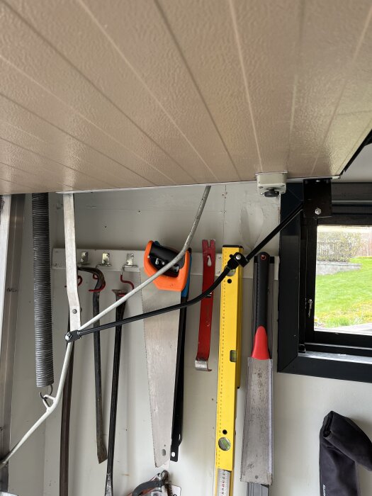 Verktyg hänger på en vägg, inklusive en såg, vattenpass och andra verktyg, med en garageport i det övre hörnet.