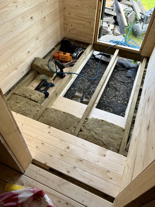 Golvisolering av stenull pågår i ett förrum, med verktyg som slagskruvdragare och material som råspontträ och stenull synliga, redo för bastuprojekt.