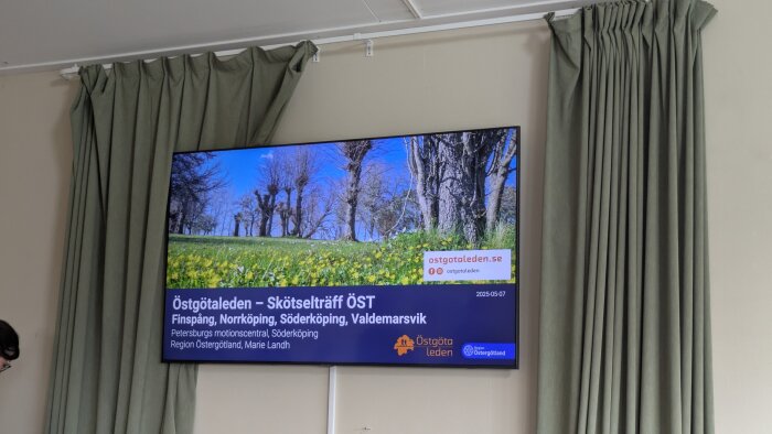 En TV-skärm visar information om Östgötaleden och en skötselträff, med en bild av träd och blommor samt kontaktinformation.
