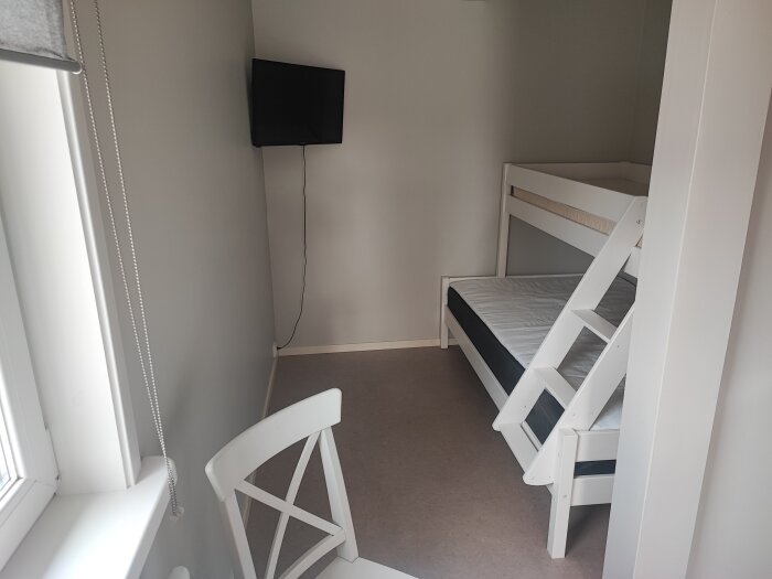 Övernattningsrum med våningssäng, väggmonterad TV och vitt bord och stol.