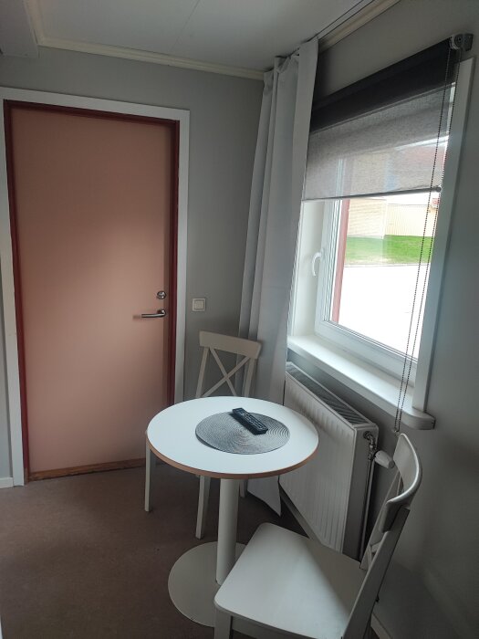 Litet rum med ett runt bord, två stolar, fönster med rullgardin och en dörr. Bordet har en fjärrkontroll på en bordstablett.