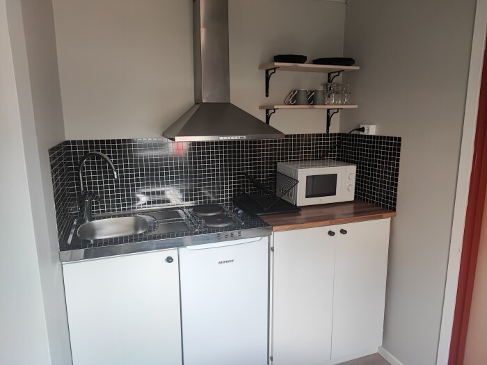 Kompakt kök med spis, diskho, mikrovågsugn och hyllor i ett övernattningsrum. Kakel i svartvitt och en saknad eluttagstäckning.