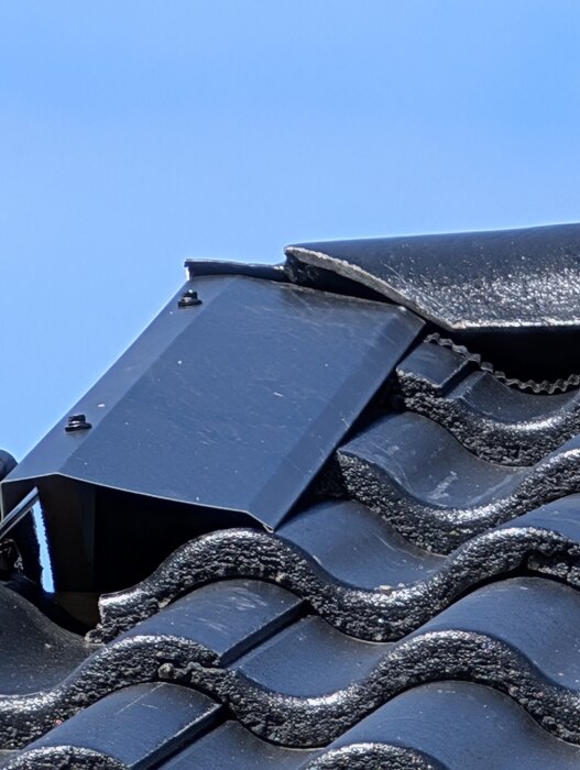 Svart vindskiveplåt över takpannor, som övergår från tegelpannor. Plåten är monterad ovanpå vågformade takpannor.
