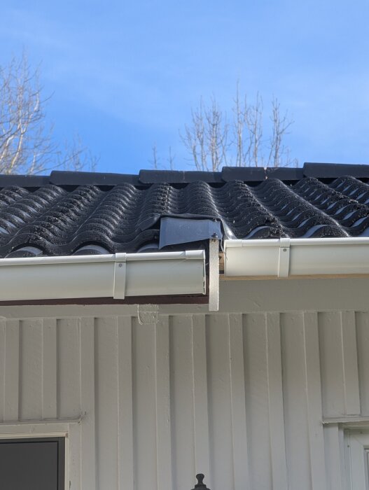 Svart tak med vågig takpanna, synliga hängrännor och vitt träpanelhus; bilden visar detaljer av taket.