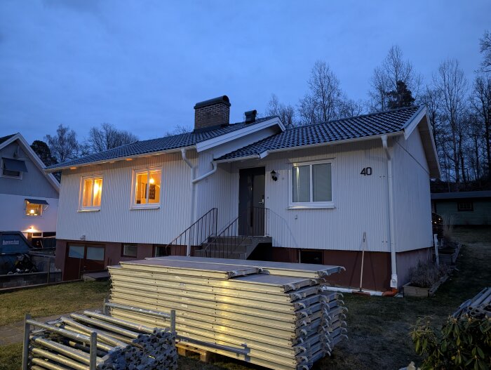 En vit enplansvilla på kvällen med stiligt lagt tak, nummer 40, och byggmaterial staplat framför huset.