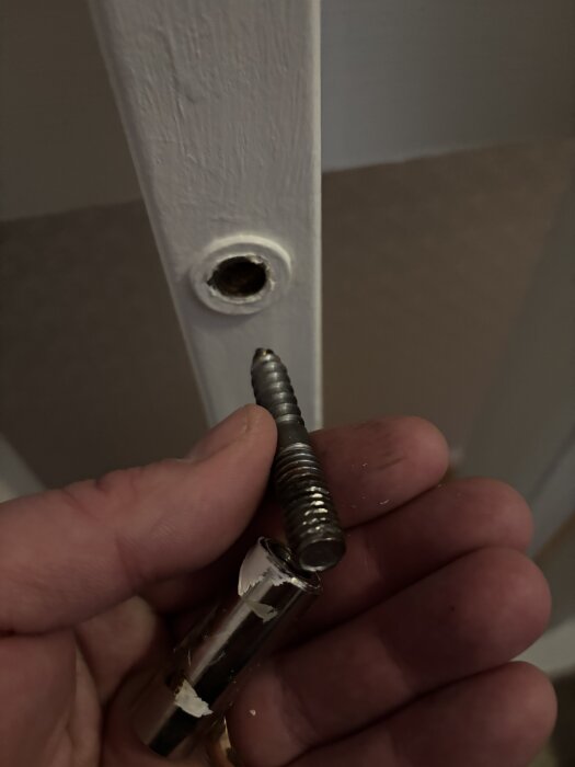 Hand håller en metallskruv framför ett borrat hål i en vitmålad yta, troligen del av ett möbelprojekt eller reparation.