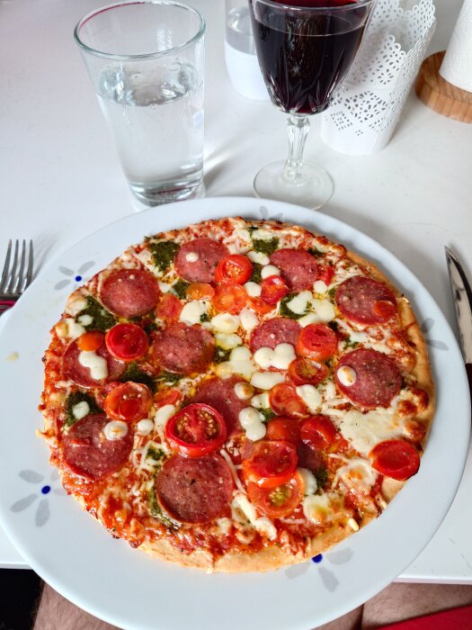 Pizza med salami, tomater, ost och pesto på en tallrik, serverad med ett glas vin och vatten på ett bord.