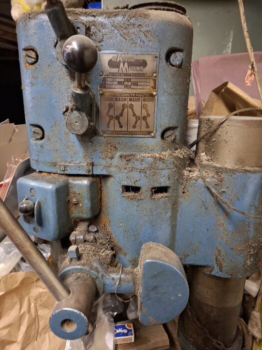 Gammal, dammig blå maskin från Arboga, Sverige, med inställningspanel och svart växelspak, omgiven av verkstadsmaterial.