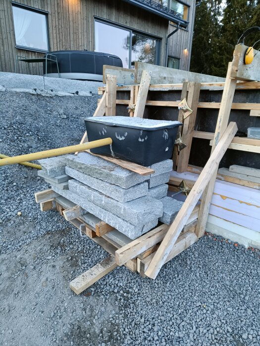 Stenplattor och en plastbalja fylld med material på en träform vid ett husbygge, använda som vikter för att stabilisera konstruktionen.