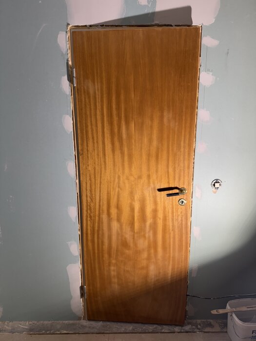 En nylackad träddörr med färgskillnader, monterad i en blågrön vägg på en byggarbetsplats. Skuggor syns på dörrens yta.