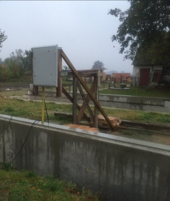 Grå betongmur på byggarbetsplats med träställning och elskåp inpå regnig dag, omgiven av grönska och äldre byggnader i bakgrunden.