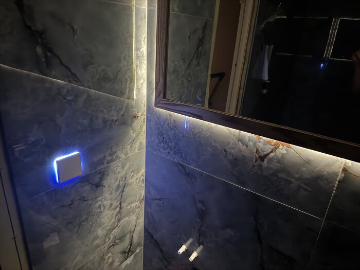 Smart switch med blått ljus integrerad i marmorerad badrumsvägg under en spegel med LED-bakgrundsbelysning.