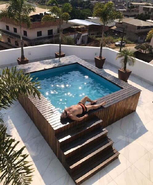 Rooftop swimming pool with wooden decking and potted palm trees; a person sunbathing beside the pool; view of surrounding buildings in the background.