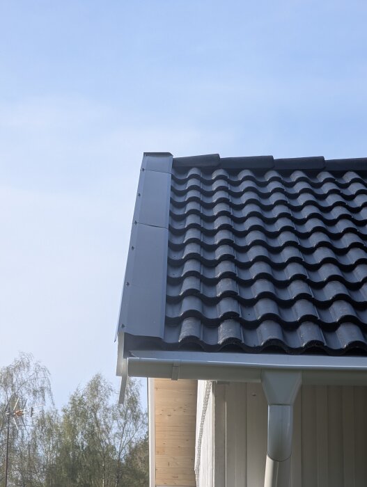 Svart tak med tegelformad plåt, där ändarna av takpannorna saknar uppvikt kant. Plåtbeslag längs kanten på taket.