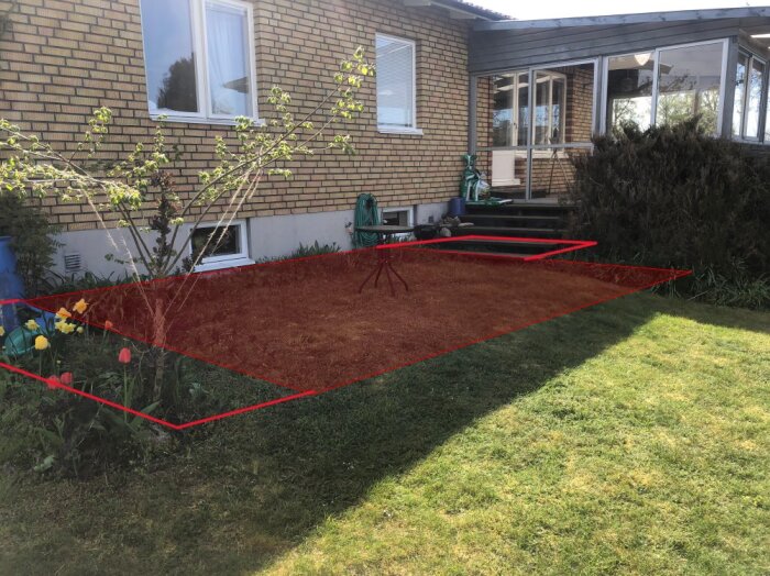 Mark med rödmarkerat område framför tegelhus där trädäck planeras, omgiven av blommor och buskar, grön gräsmatta synlig.