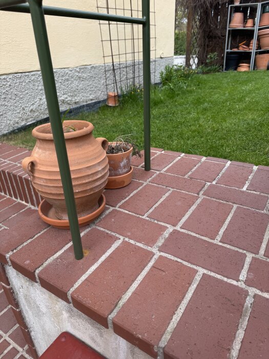 Punainen tiiliterassi, jossa on kaksi kukkaruukkua ja vihreä metallikaide. Taustalla nurmikko ja seinää vasten metallisäleikkö.