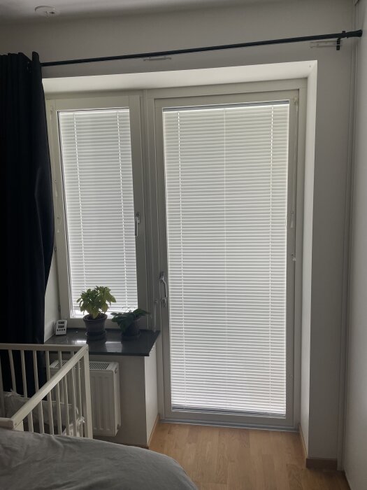 Rummet med vita persienner och mörkläggningsgardiner, ett fönster och en altandörr, samt en fönsterbräda med två växter.