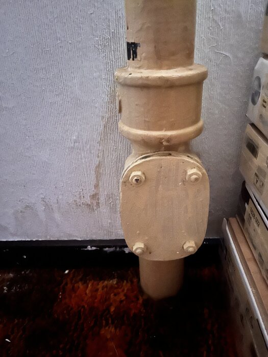 Avloppsrör i källare med skarv och renslucka, omgivna av kartonger och fuktfläckar på väggen.