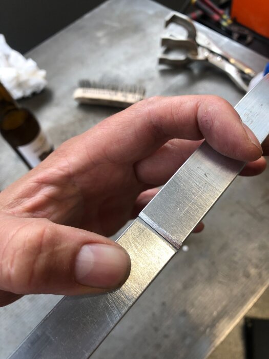 Hand som håller en böjd aluminiumstång med synlig svetsfog, verktyg och borste i bakgrunden på metallbord.