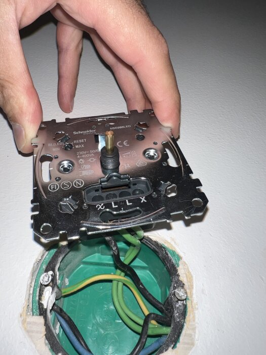 Hand håller en Schenider-dimmer ovanför en grön kopplingsdosa med exponerade kablar i blått, svart och gulgrönt.