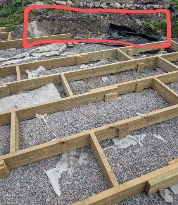 Bygge av en altan på ojämn, bergig mark med träramverk, synlig stenkant, samt en markerad del med ogräs och svart jordduk i bakgrunden.