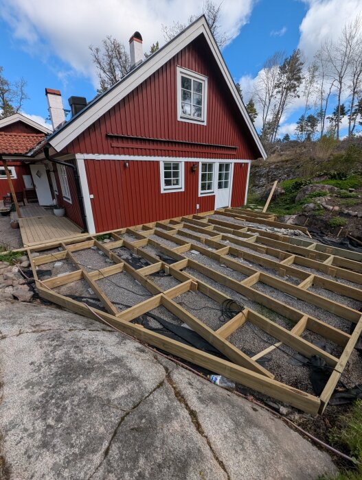 Träram för altanbygge vid en röd stuga på en ojämn och bergig tomt, med sten och grus på marken.