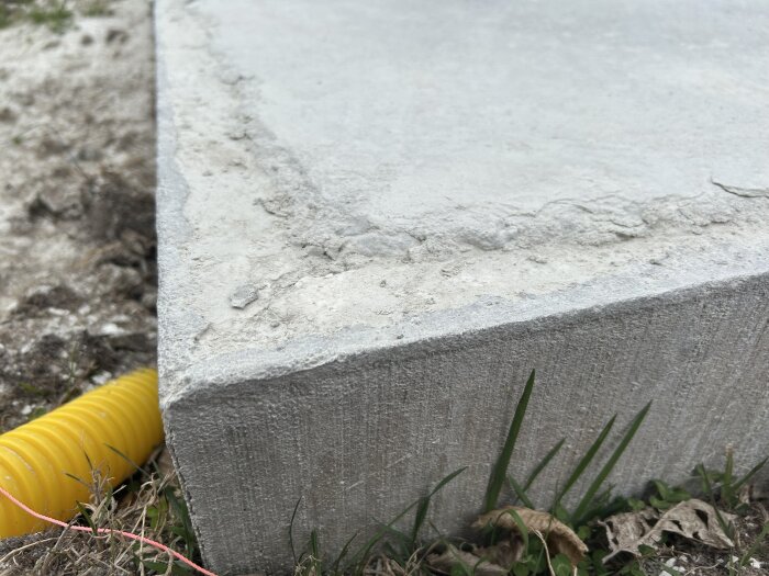 Närbild av ett gjutet betongfundament i ett hörn, med ojämna ytor och gräs vid sidan; synlig gul dräneringsslang intill.