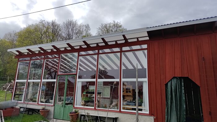 Ett rött hus med stora fönsterpartier, genom vilka växter och trädgårdsredskap syns. Taket har snören som fungerande öppnare.