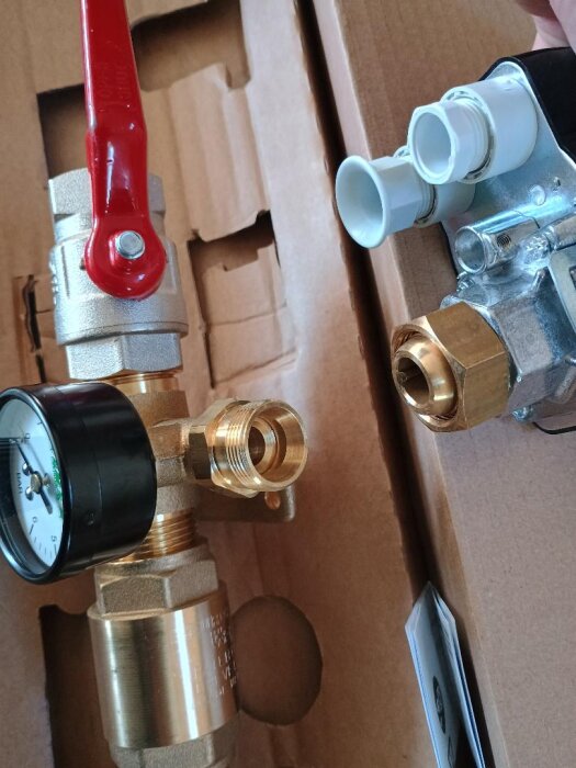 VVS-komponenter på en kartong, inklusive ett kopplingsstycke med röd ventil och tryckmätare, samt rördelar i plast och metallkopplingar.