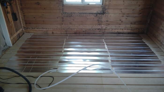 Renoverat rum med träväggar och golv med silverfärgade spår för rörläggning, visar installation av värmesystem under golv.