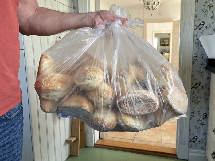 En hand håller en genomskinlig plastpåse fylld med bröd i ett kök med mönstrade tapeter och trägolv i bakgrunden.