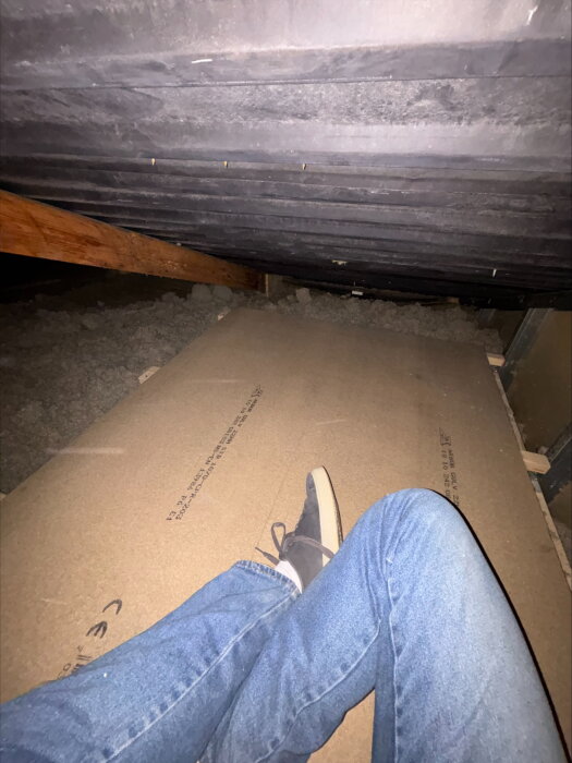 Person i jeans och sneakers ligger på vinden under ett plåttak med synliga takstolar och isolering.