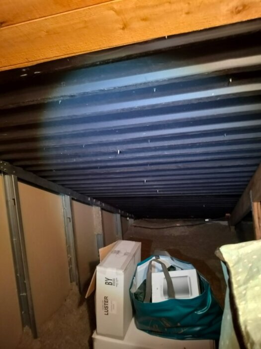 Oinredd vind med plåttak och brist på ventilation; synliga kartonger och påsar ligger under taket.