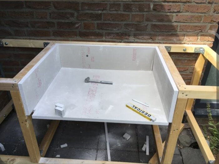 Inbyggnad av grill i utekök med cementboard installerad på träram, omgiven av tegelvägg. Inkluderar verktyg och en vattenpass på ytan.