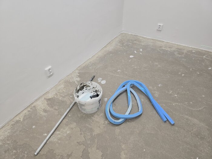 Byggarbetsplats med betonggolv, en hink med verktyg och blå avstängningslister, i ett rum med vita väggar och eluttag.