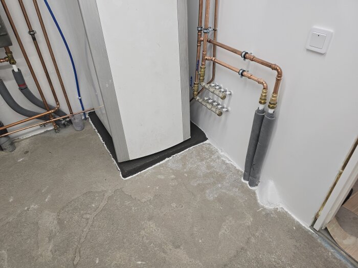 Golvyta i maskinrum med synliga betonggolv, kopparrör, och en stor vit enhet med svart bas. Röret har stöd av vita avstängningslister.