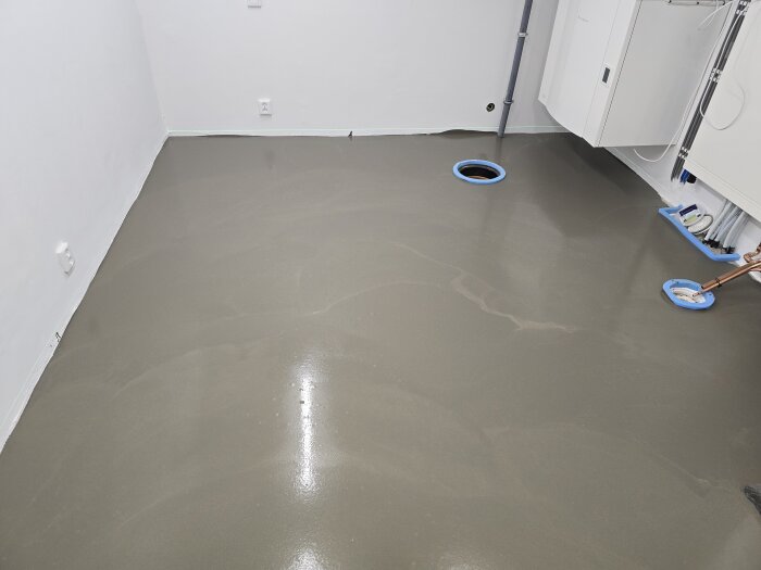 Nytt flytspacklat golv i maskinrummet med två synliga rörinspektioner i blå skyddsringar. Vitmålade väggar med eluttag syns också.