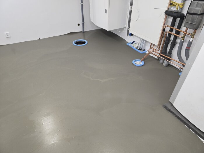 Golvyta i maskinrum med nypålagt flytspackel, synliga avlopps- och vattenrör samt vägguttag, redo för plattsättning.