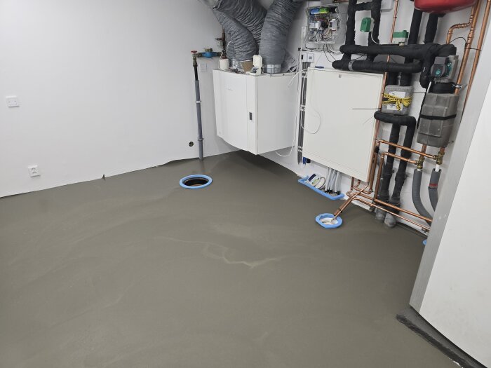 Maskinrum med nylagt flytspackel, rör och elinstallationer synliga. Inspektionslucka och golvdränering markerade med blå cirklar.
