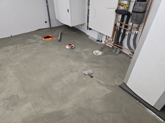 Golv med nylagt flytspackel i maskinrum, synliga rör och verktyg; förberett för plattsättning, syns nära avloppet och vägginstallationer.