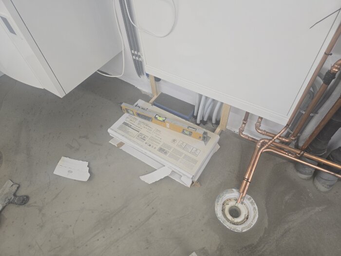 Byggmaterial och kopparrör i ett maskinrum förbereds för plattsättning; papplåda med klinkers och verktyg syns på betonggolvet.