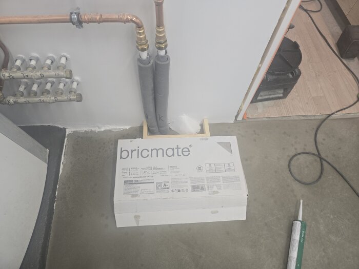 En paket med Bricmate-plattor står på ett betonggolv, omgiven av rör och verktyg i ett pågående renoveringsprojekt.