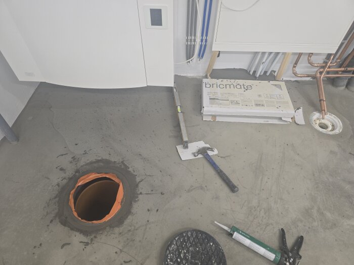 Golvet i ett maskinrum med en öppen inspektionslucka, verktyg och ett kakelpaket synliga. Golvets yta förbereds för plattsättning.