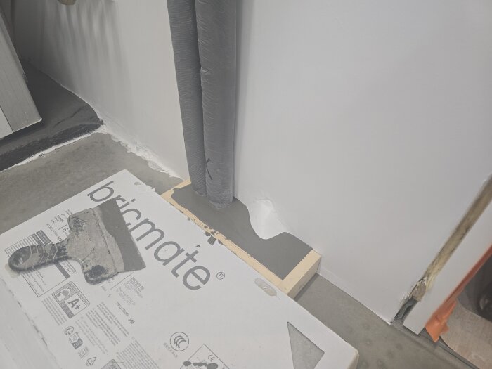 En spackelspade ligger på en kartong märkt "bricmate" vid en vägg, där flytspackel applicerats nära rör och en inspektionslucka.