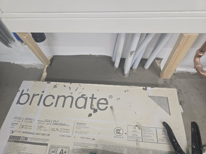 Bild av ett maskinrum under renovering med ett paket Bricmate klinkerplattor på golvet, betongunderlag, trästöd och synliga rör.